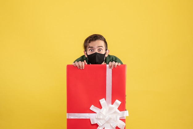 Free photo front view wide-eyed man with mask hiding behind big giftbox on yellow