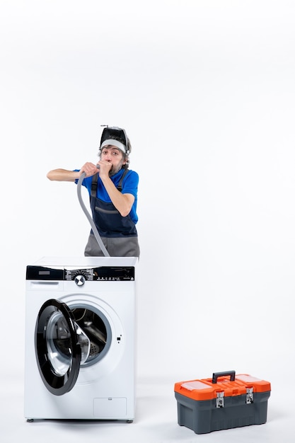 Front view of wide-eyed repairman in uniform standing behind washing machine blowing out pipe on white isolated wall