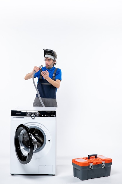 Front view of wide-eyed repairman in uniform standing behind washing machine checking pipe on white wall