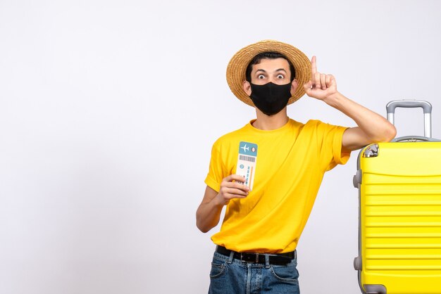 Front view wide-eyed young man with straw hat standing near yellow suitcase holding travel ticket pointing finger up