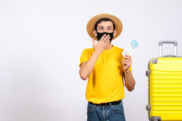 Front view wide-eyed young man with straw hat standing near yellow suitcase holding travel ticket