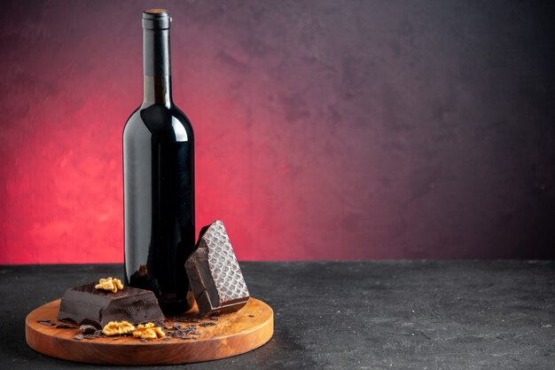 Вид спереди бутылка вина кусочки грецкого ореха темного шоколада на деревянной доске на красном фоне