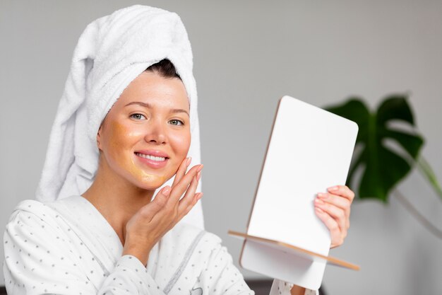Вид спереди женщины в халате, применяющей уход за кожей с полотенцем на голове