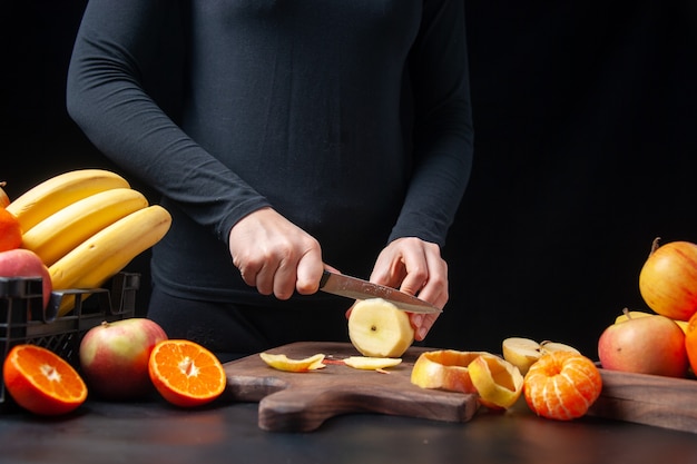 Front view of woman chopping fresh apple on wooden board fruits in wooden tray and plastic box on table