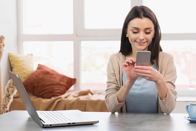 Front view of woman at desk with laptop holding smartphone