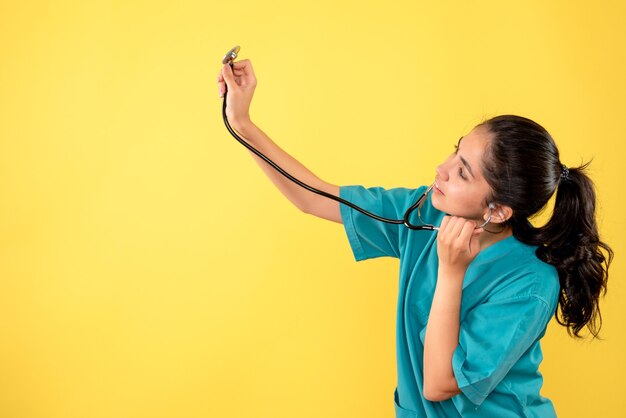 Front view of woman doctor in uniform raising stethoscope on yellow wall