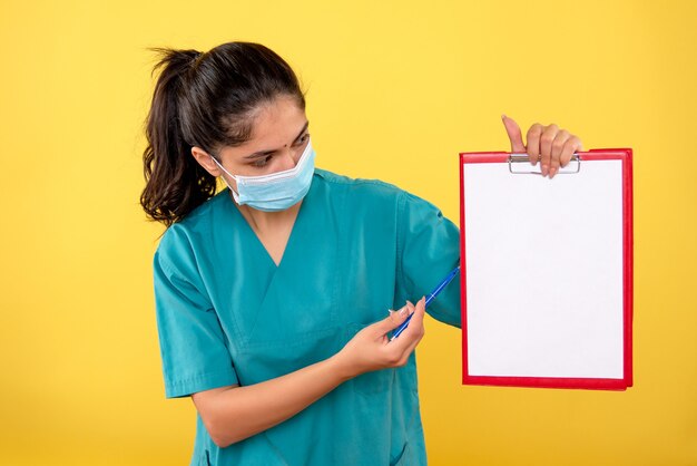 Вид спереди женщины-врача в униформе, показывающей что-то на бумаге на желтой стене