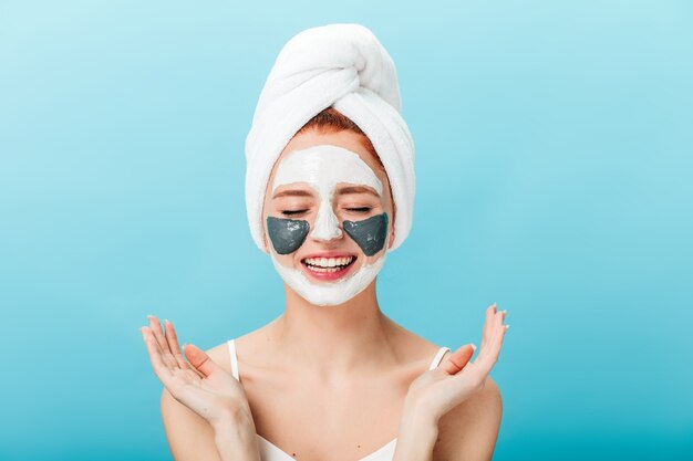 Front view of woman doing spa treatment with closed eyes. Studio shot of charming girl with face mask standing on blue background.