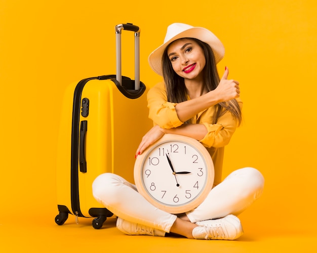 Front view of woman holding clock and posing next to luggage