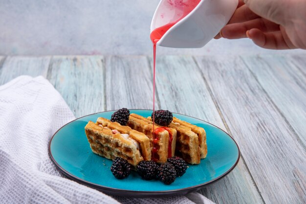 Front view of woman pours syrup sweet waffles with blackberries on a blue plate with a white towel on a gray surface