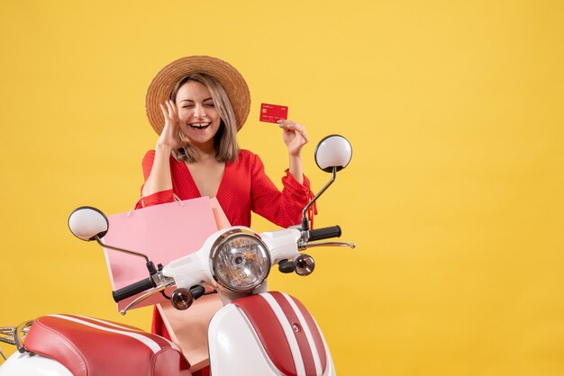 Front view of woman in red dress on moped holding shopping bags and card