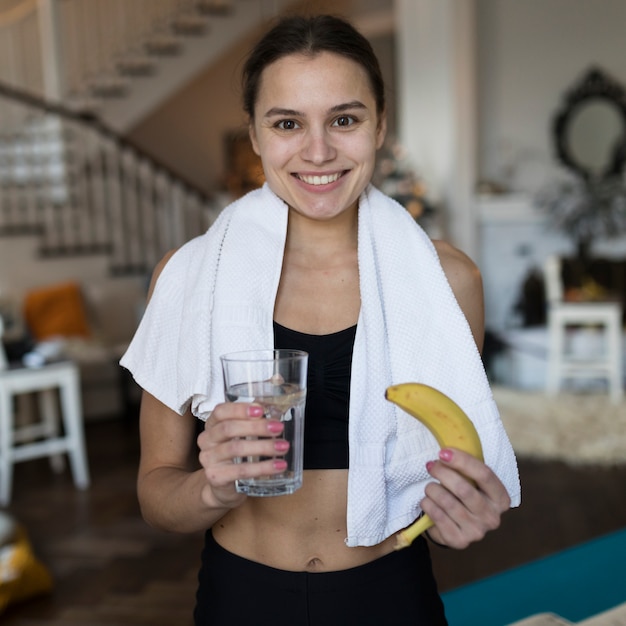 Вид спереди женщины в спортивной одежде, улыбаясь, держа банан и стакан воды