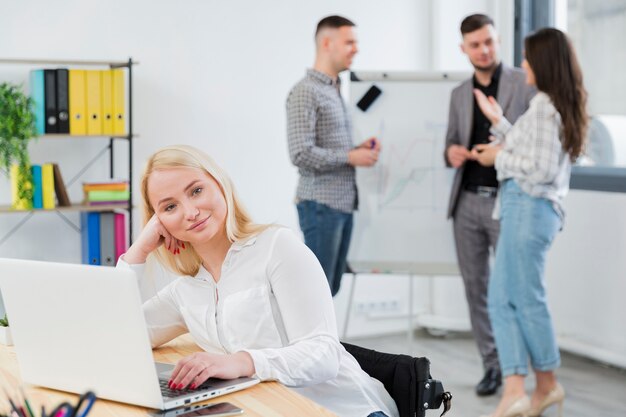 Front view of woman in wheelchair posing at work while colleagues converse