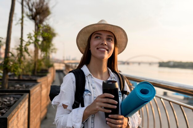 Front view of woman with backpack and hat holding thermos while traveling