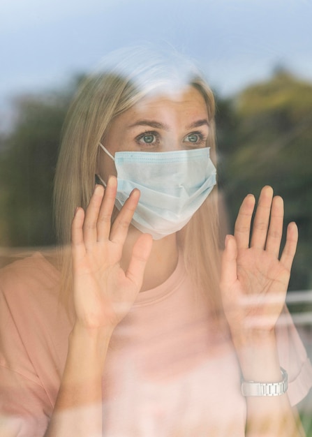 Foto gratuita vista frontale della donna con mascherina medica a casa toccando la finestra durante la pandemia