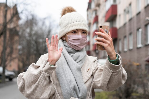 スマートフォンで手を振っている医療マスクを持つ女性の正面図