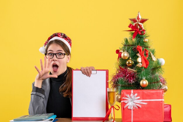 Front view wondered girl with xmas hat sitting at the table opening her mouth xmas tree and gifts cocktail