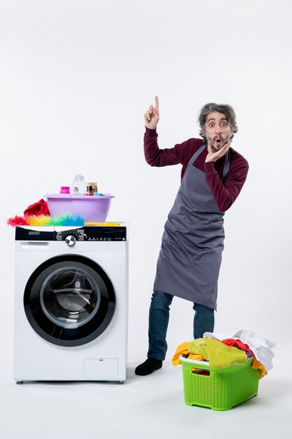 Front view wondered housekeeper man standing near washer laundry basket on white wall