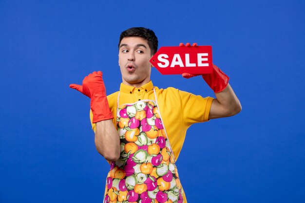 Вид спереди удивленного домработника в красных сливных перчатках, держащего знак продажи, указывающий налево на синей стене