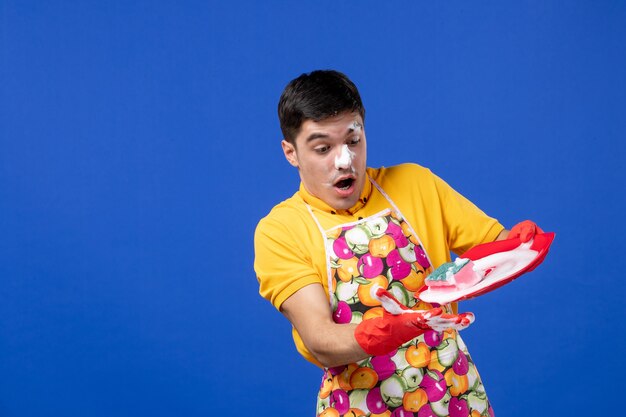 Front view of wondered housekeeper with foam on his face holding plate with foam on blue wall