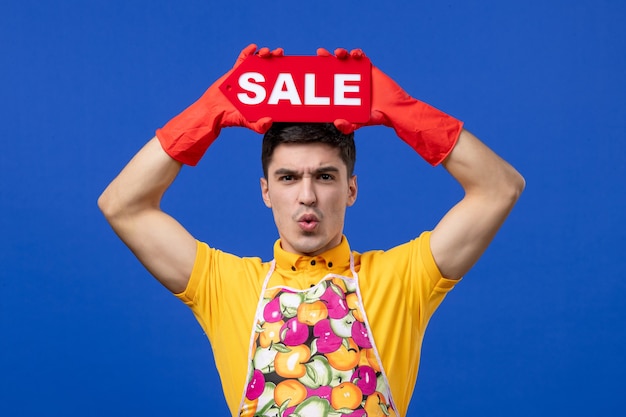Free photo front view of wondered male housekeeper in apron raising sale sign over his head on blue wall