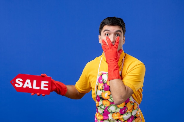 Free photo front view of wondered male housekeeper in yellow t-shirt holding sale sign putting hand on his face on blue wall