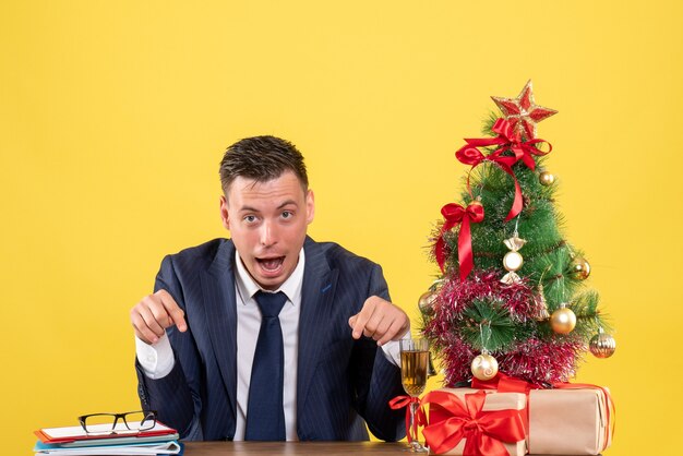 クリスマスツリーと黄色の贈り物の近くのテーブルに座っているテーブルを指している不思議な男の指の正面図
