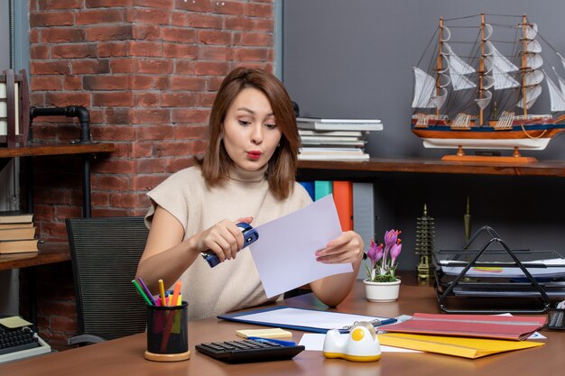 Front view of wondered woman using stapler sitting at office