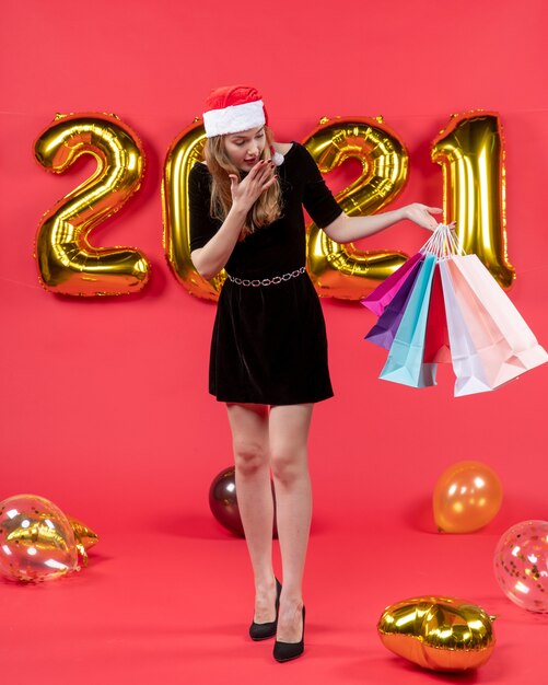 Front view wondered young lady in black dress holding shopping bags balloons on red
