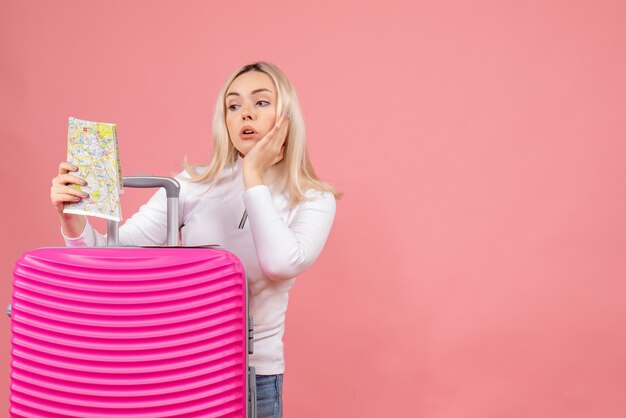 Front view wondered young lady standing behind pink suitcase looking at map