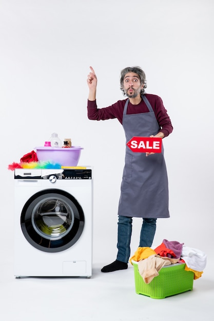 Free photo front view wondered young man in apron holding up sale sign standing near washer on white wall