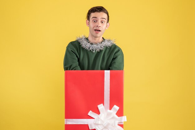 Front view wondered young man standing behind big giftbox on yellow 