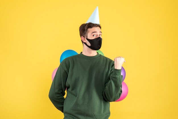 Front view wondered young man with party cap and colorful balloons standing on yellow 