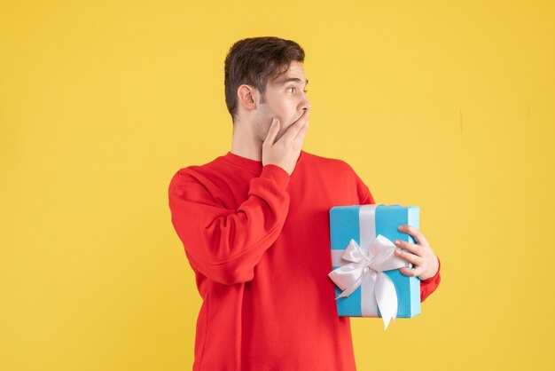 Front view wondered young man with red sweater holding blue gift box on yellow 
