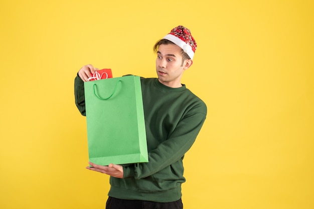 Front view wondered young man with santa hat holding green shopping bag and gift standing on yellow 