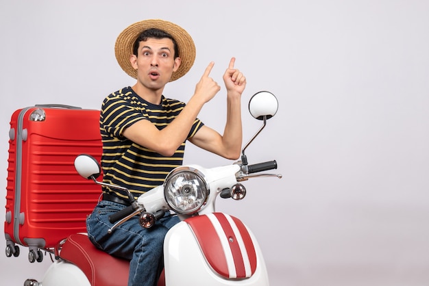 Front view of wondered young man with straw hat on moped pointing at behind