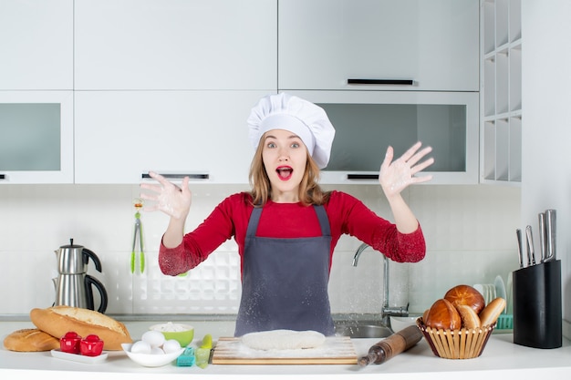 Front view wondered young woman in cook hat and apron raising hands with flour in the kitchen