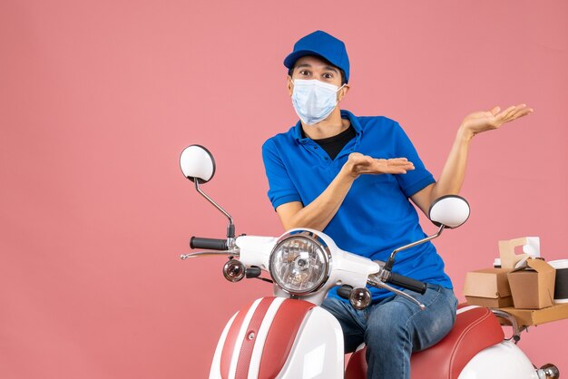 Front view of wondering courier man in medical mask wearing hat sitting on scooter on pastel peach background