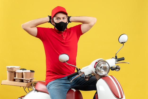 Front view of wondering courier man wearing red blouse and hat gloves in medical mask delivering order sitting on scooter