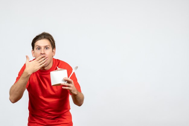 Вид спереди удивленного молодого парня в красной блузке, держащего бумажную коробку и ложку на белом фоне