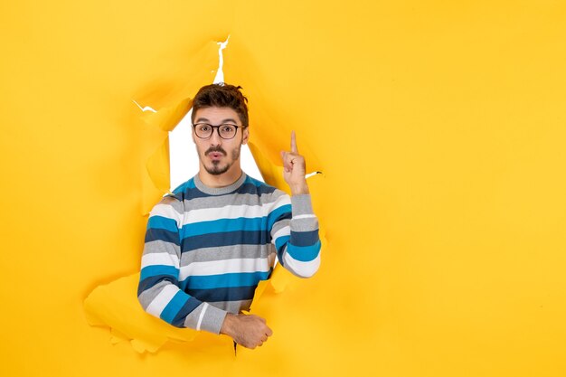 Front view wondering young man pointing finger up looking through ripped paper yellow wall