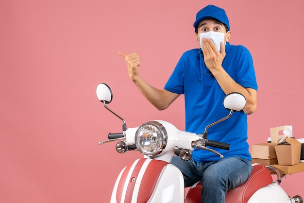 Front view of worried delivery guy in medical mask wearing hat sitting on scooter on pastel peach background