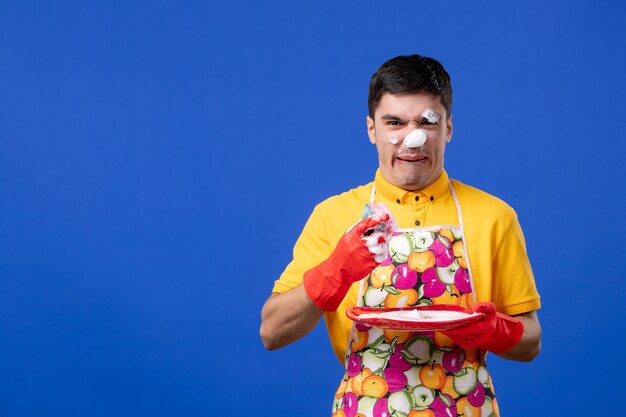 Front view of worried housekeeper with foam on his face washing plate on blue wall