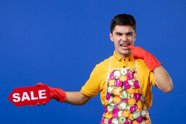 Front view of worried male housekeeper in yellow t-shirt holding sale sign biting his finger on blue wall