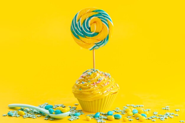 A front view yellow cake with lollipop on top along with colored candies on yellow