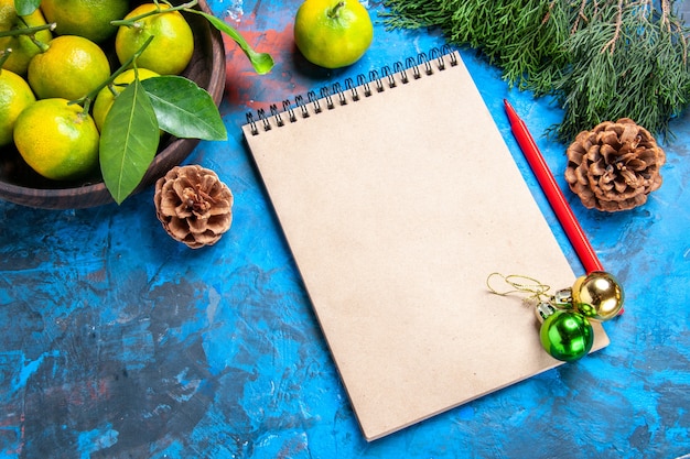 Front view yellow tangerines with leaves in wooden bowl a notebook red pencil xmas details on blue background