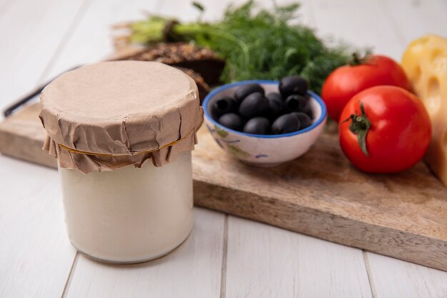 Йогурт вид спереди в банке с оливками, помидорами, ломтиками черного хлеба и укропом на подставке на белом фоне