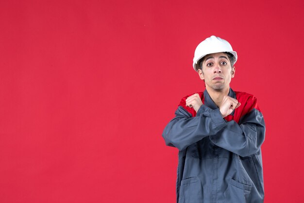 Front view of young architect in uniform with hard hat and making stop gesture on isolated red wall