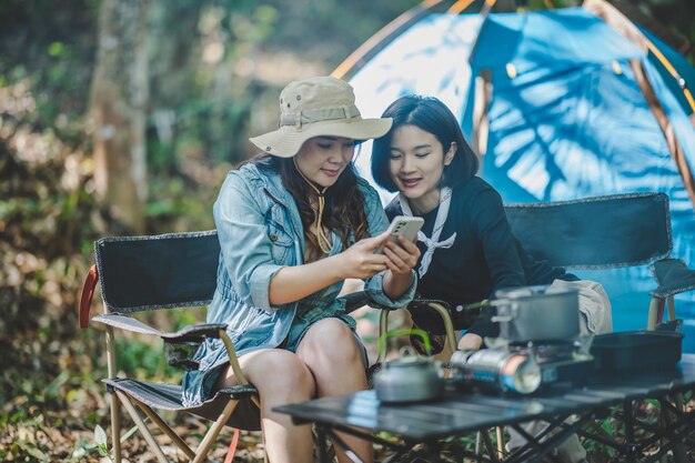 正面図テントの前に座っている若いアジアのきれいな女性と彼女のガールフレンドは携帯電話を使用して一緒に幸せで森のキャンプ中に写真を撮ります