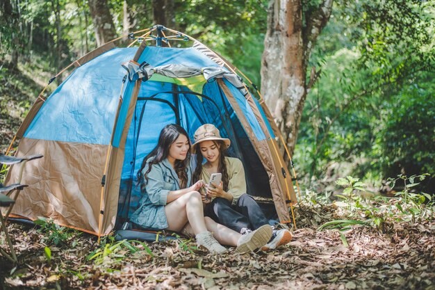 전면 보기 텐트 앞에 앉아 있는 젊은 아시아 미녀와 그녀의 여자친구는 휴대전화를 사용하여 함께 숲에서 야영하는 동안 행복과 함께 사진을 찍는다